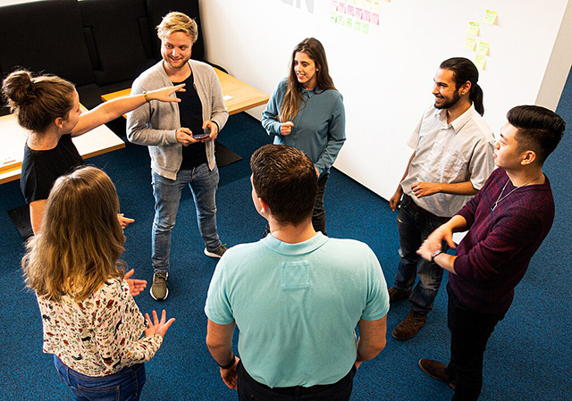 Sieben Personen stehen locker in einem Kreis und besprechen sich. Im Hintergrund sieht man eine Pinnwand mit bunten Klebezetteln. Sieben Personen stehen locker in einem Kreis und besprechen sich. Im Hintergrund sieht man eine Pinnwand mit bunten Klebezetteln.