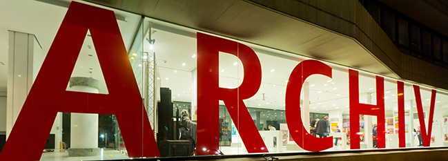 Das Bild zeigt eine lange und hell erleuchtete Fensterfront. Darauf steht in großen, roten Buchstaben das Wort "Archiv". Das Bild zeigt eine lange und hell erleuchtete Fensterfront. Darauf steht in großen, roten Buchstaben das Wort "Archiv".