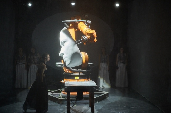 Eine Frau tanzt vor einem überdimensioniertem Robotergesicht.